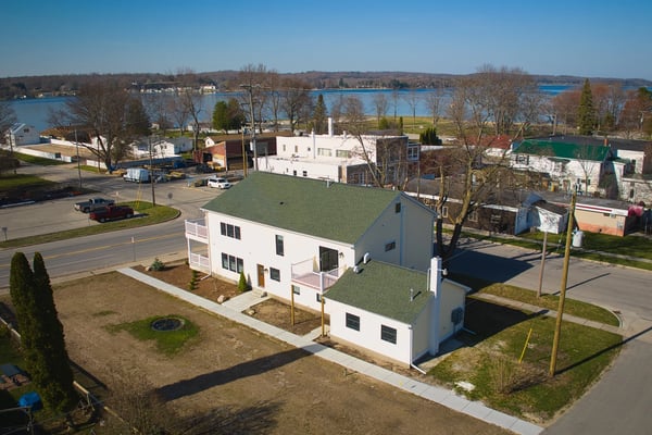 Gray shingled beach cottage with deck, white patio furniture, and sandy yard facing water.