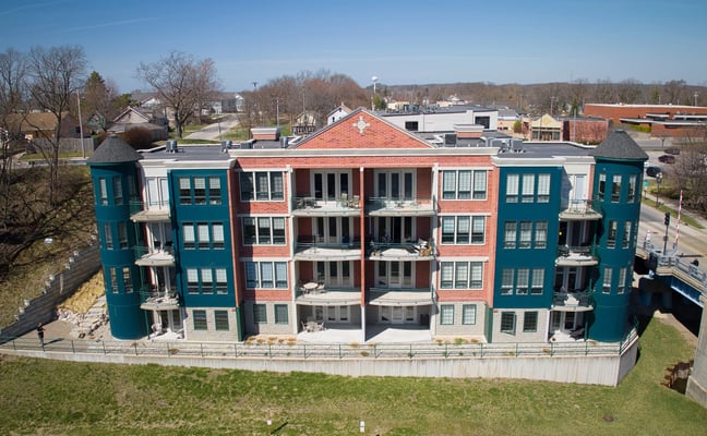 Multi-unit residential building with teal corner towers and red-brick center section, white balconies, overlooking green lawn and small town.